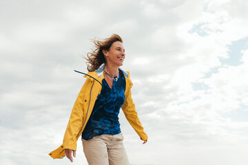 happy woman with blowing hair in a yellow raincoat looking at the ocean on a windy day