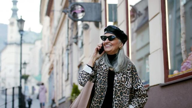 Senior chic woman with sunglasses standing outdoors, using smartphone.