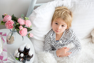 Sick little blond toddler boy, lying in bed with fever, hugging little fluffy toy, medicine next to him