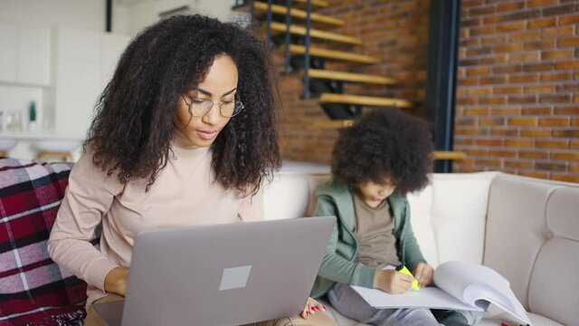 Single mom. Portrait of mixed race girl with long curly hair working with laptop at home while her preschooler son draws with felt-tip pens in a notebook