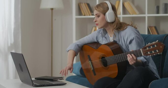 female musician is playing guitar at home, listening to music by headphones, spending time alone at weekend