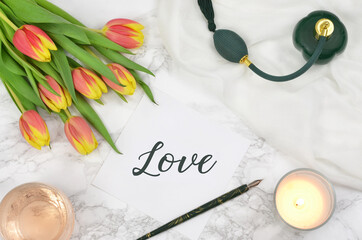 Inscription "love", flowers, perfumes, candle, pen, top view on a marble background, flat lay on light textured stone table, trendy background.