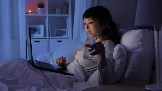 technology, internet, communication and people concept - young asian woman with laptop computer lying in bed and drinking water at home at night