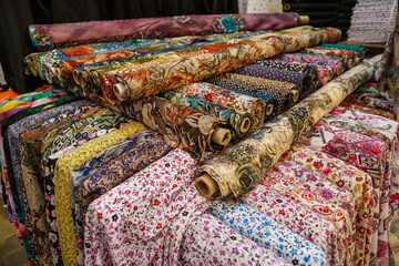 Fabrics with different designs and colors stacked outside a market.