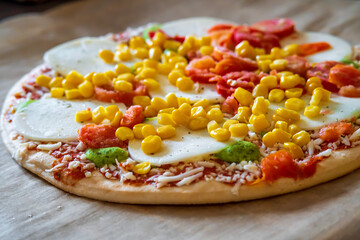 Colorful raw pizza before baking with mozzarella cheese, corn, pesto and cherry tomatoes. Frozen, unbaked, gluten free pizza placed on a baking sheet before putting it in the oven. Side view.