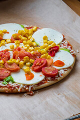 Colorful raw pizza before baking with mozzarella cheese, corn, pesto and cherry tomatoes. Frozen, unbaked, gluten free pizza placed on a baking sheet before putting it in the oven. Side view.