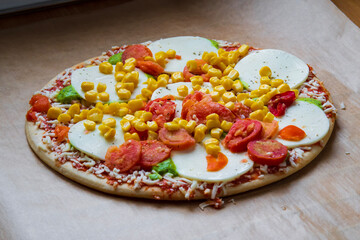 Colorful raw pizza before baking with mozzarella cheese, corn, pesto and cherry tomatoes. Frozen, unbaked, gluten free pizza placed on a baking sheet before putting it in the oven. Side view.