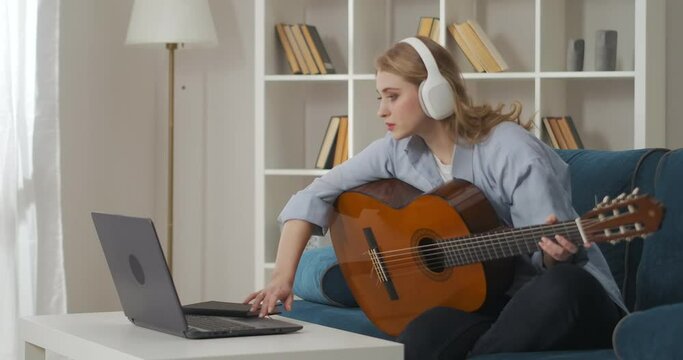 female musician is opening sheet music on laptop display and playing song on guitar, relaxing at home at weekend