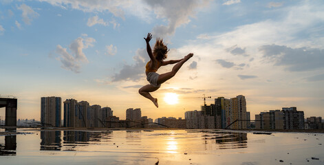 Female flexible dancer jumping during sunset on cityscape background with reflection in the water. Concept of freedom and happiness 