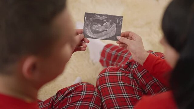 Shooting over shoulder of happy Caucasian couple admiring ultrasound picture sitting at home indoors. Happy man and woman holding photo of fetus looking at each other smiling. Happiness and pregnancy.