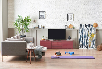 Sportive and training decoration interior room, nobody, television unit, grey sofa and blanket. Purple mat and blue dumbbell on the parquet, white brick wall background.