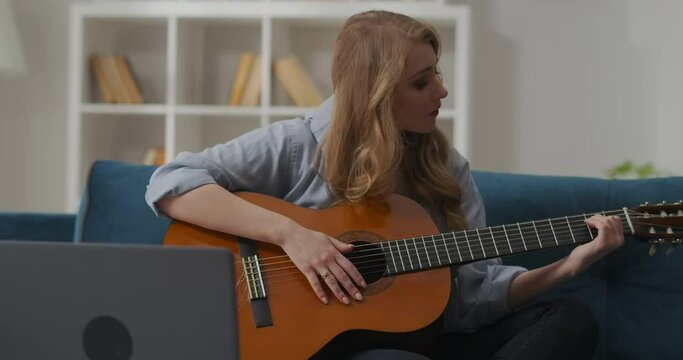young female beginner musician is learning accords on guitar, playing on strings, sitting on couch in apartment