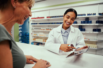 Cheerful young woman wearing labcoat working in pharmacy showing screen to female customer on...