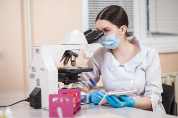 A young female laboratory assistant in a white uniform works in his office behind a microscope on the background of a medical clinic. Concept medicine, biology, research