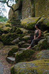 Beautiful young caucasian woman sitting with a green dress on a staircase in the outdoors full of rocks and trees with moss