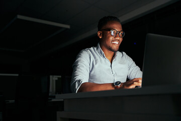 Smiling african young businessman working overtime in the office to finish the project within the...