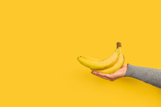 Mano femenina sosteniendo pl&aacute;tanos sobre un fondo amarillo liso y aislado. Vista de frente. Copy space. Concepto de nutrici&oacute;n, salud.