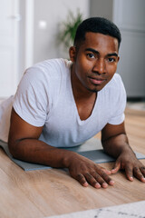 Close-up view of handsome African-American man exercising in horizontal plank position on floor during working out at bright domestic room. Concept of sport training at home gym.