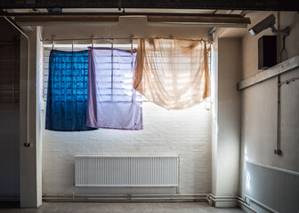 Coloured prisoner bedsheets hanging as curtains over prison bars sunlight in window of high security run down immigration detention centre in England with CCTV security camera monitoring the room
