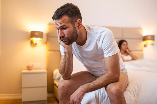 Young Sad Man Sitting On Bed After Quarrel With Wife. Hotel, Travel, Relationships, And Sexual Problems Concept - Upset Man Sitting On The Bed With Woman On The Back