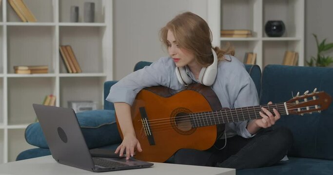woman is looking for information how to play guitar in internet on laptop, holding guitar and plucking strings, self-education