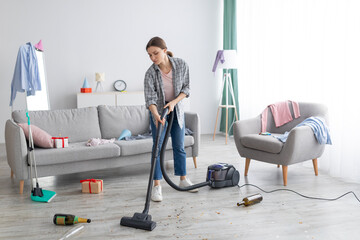 Unhappy young woman vacuuming messy apartment after party, cleaning up chaos after holiday celebration, copy space
