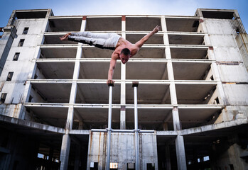 Flexible acrobat doing handstand on the industrial construction background with columns on sides....