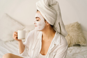 Beautiful woman applying facial mask and drinking tea in bed