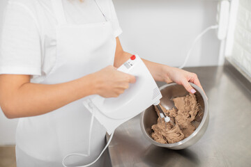 Hands of female professional confectioner holding the hand mixer, immersed in the chocolate butter cream. Whipping the cream for ganache. Baking process stages