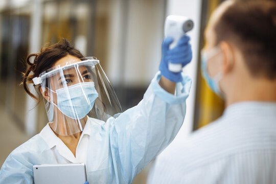 Nurse Measures Temperature Of A Patient Hospital Visitor With A Non Contact Thermometer. People Wearing Protective Medical Masks. Coronavirus Prevention And Healthcare Concept.