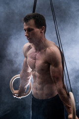 Fototapeta na wymiar Muscular build man doing calisthenics on gymnastics rings indoor on black, smoked background. concept of healthy lifestyle and power.