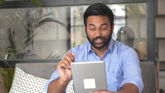Smiling Indian businessman, worker talking and discussing on video conference online using tablet at home office. Male professional explaining project with gestures at remote workplace.