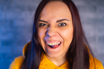 Portrait of young woman with grimacing face. Close up of crazy female in yellow hoodie monkeying on gray brick background. - obrazy, fototapety, plakaty