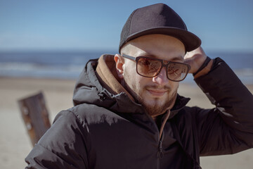 Young stylish hipster in a black cap, jacket walking and relaxing on a beach.Relaxed and cheerful. Outdoor portrait of happy young caucasian man near the sea.