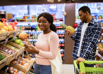 Happy black lady checking shopping list on mobile phone while choosing fruits with her boyfriend at supermarket