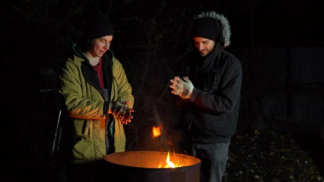 Two homeless young men bask in the fire. Men stand near a barrel of fire at night.