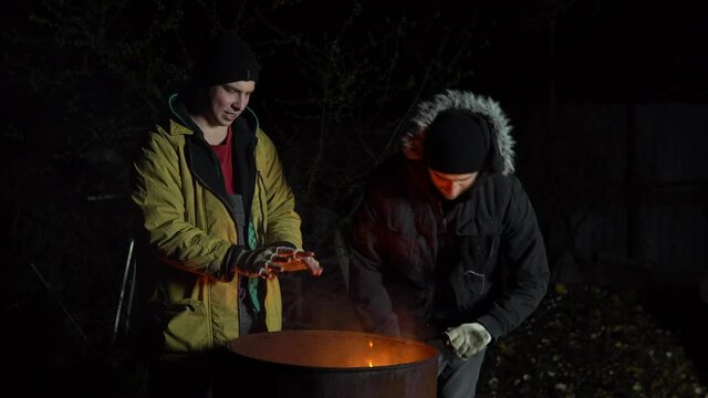 Two homeless young men bask in the fire. Men stand near a barrel of fire at night.