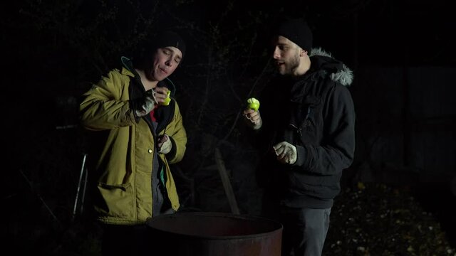 Two homeless young men are frying apples on a fire. Men stand at night near a barrel of fire and bask.
