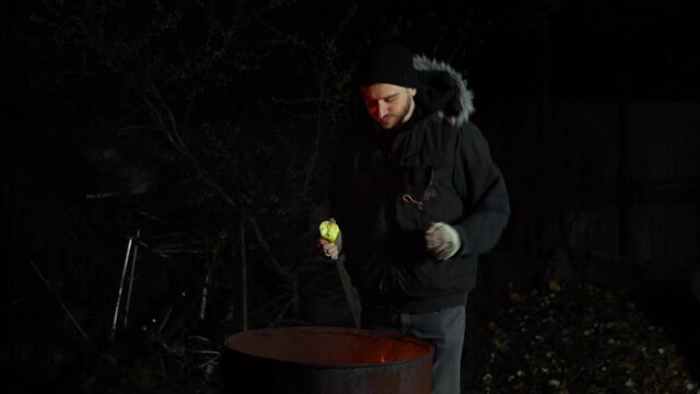 A homeless young man stands by the fire and eats an apple. A man stands at night near a barrel of fire and bask.