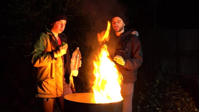 Two homeless young men stand by the fire and eat apples. Men stand at night near a barrel of fire and bask.