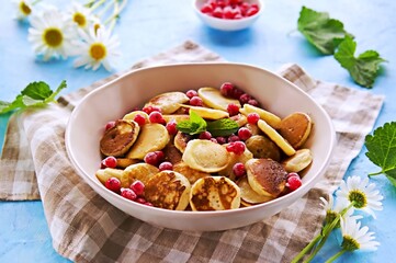Mini pancakes with red currants in a beige bowl on a blue concrete background.