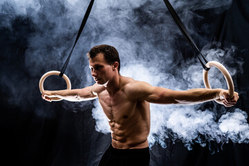Muscular build man doing calisthenics on gymnastics rings indoor on black, smoked background. concept of motivation, desire and passion - obrazy, fototapety, plakaty