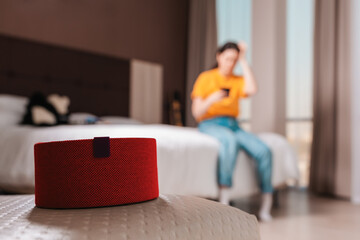 Obraz premium Red portable smart speaker close-up. In the background, a woman is sitting on a bed with a phone, in a blur. The concept of modern gadgets
