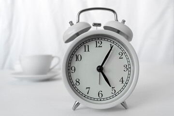 The white alarm clock is on the table.Behind it is a white cup of coffee on a light background. The image creates an atmosphere of the morning, early time of day