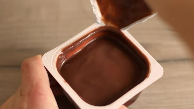 Man hand opens plastic container with chocolate mousse or pudding on wooden table in the kitchen