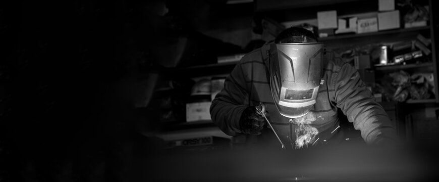 An adult male works in an old workshop. He holds a welding machine in his hand and welds the metal structure. Copy space. Black and white.