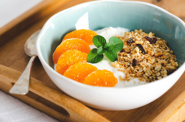 Plain yoghurt with tangerine segments, granola, and mint leaves in a white and light blue bowl, with a spoon, on a wooden tray. 