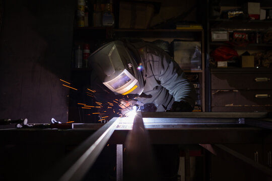 An adult male works in an old workshop. He holds a welding machine in his hand and welds the metal structure.