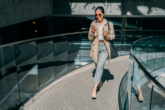 front view full length of happy asian japanese businesswoman walking on city modern building using smartphone online texting message. elegant office lady in high heel shoes with cup of morning coffee