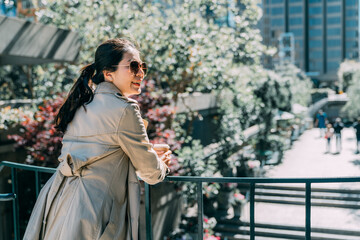 Obraz na płótnie Canvas back view elegant young asian japanese woman standing outdoor modern office building in city and holding take away coffee. smiling office lady employee leaning on railing looking side on sunny day.